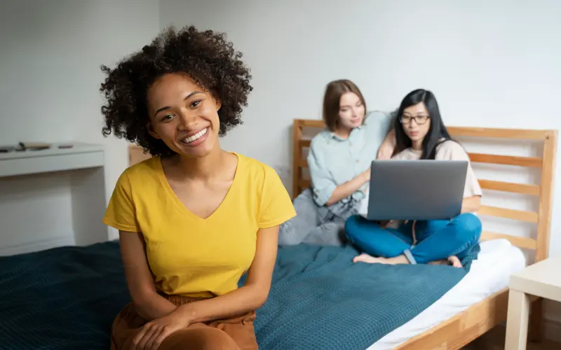 Smiling student sitting on her bed in a shared room, roommates on a laptop behind her